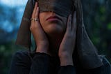 Image of a woman blindfolding her eyes with a brown scarf. She’s holding the scarf and her face.