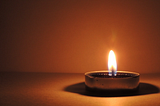 A single candle burning in darkness, its flame casting a soft, warm glow against a muted background, symbolizing quiet reflection, hope, and vigilance.