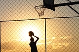 Person holding basketball under basketball goal as sun sets.