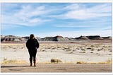 A plus size woman with midlength brown hair is standing on an empty road, dressed in all black and brown knee boots, facing away from the camera, toward a barren desert landscape with gray and red hills in the distance against a bright blue sky.