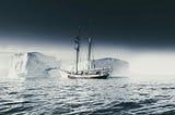 A small sailing ship passes a towering iceberg across calm polar waters beneath a dark, expansive sky, evoking uncertainty, exploration, and navigation.