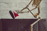 An adorable skeleton sits on a ledge with a red cap on, tying their red high-top converse sneakers