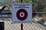 Sign on a fence with a bulls-eye that says “touch the bulls-eye or your lap doesn’t count.”
