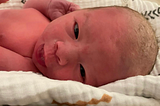 A pink newborn baby stares into the camera with vernix still in hair.