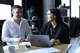 Two people in an office sitting and smiling at one another in front of a computer.