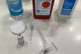 A bathroom counter with a variety of medical supplies, including a syringe, needles, sharps container, alcohol, and a vial of estradiol.