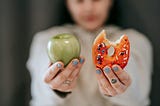 A blurred, out of focus person holding up an apple on the right and a glazed donut with a few sprinkles with a bite taken out of it on the left.
