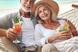 An older couple relaxing in a hammock on a beach drinking cocktails.