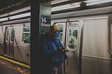 Man in blue hoodie using a smartphone in 14 Street station with a train nearby.