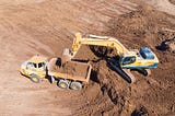 An articulated dump truck is to the left in the photo and positioned lower than the hydraulic excavator which is loading it from the right and above. The excavator is emptying a load of dirt from its bucket into the truck, which already is holding some dirt.