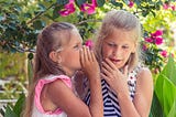 2 girls whispering. Being ‘2 faced’ helps society function better.