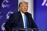 Donald Trump stands at a podium and delivers a speech. He wears a dark suit, white shirt, and blue tie with white stripes.