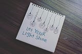 Picture of a notebook on a wooden table. There are drawn lightbulbs on the notebook and the words Let Your Light Shine.