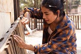 Young woman wearing a loose checked shirt, pencil stuck in her hair, hammering a nail into a bedframe.