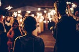 Several wedding guests with sparklers lighting up the night, a young man and woman in the foreground facing away from the camera, a young woman looking back at the couple.