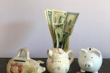 Three piggy banks on a table, with piles of coins and bills stuck in one bank.