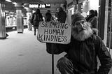 A man holding a sign in a public place that reads “Seeking human kindness.”