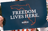 Photo of a red, white and blue campaign poster for Ron DeSantis for Governor of Florida that says “Freedom Lives Here” over an outline of the State of Florida