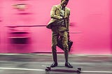 Professional Black man wearing tan suit, carrying briefcase and riding a skateboard down a street while looking at his watch.