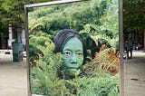 A framed large photograph of an Asian woman painted green, hiding among foliage. Her eyes are bloodshot and her hair is black. This photo was displayed as part of an Art exhibition in Central London.