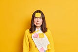 A serious female has a post-it note stuck on her forehead and looks as if she is thinking about a memoir. Bright yellow background.