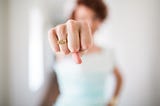 I woman stands with her fist in the camera. The fist is in focus and the woman is out of focus in the background. The woman wears a gold ring on her ring finger that says “I am bad ass”