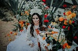 A Woman in Catrina Makeup Sitting beside an Altar with Food and Flowers