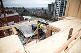 Photograph of a construction worker working on a timber building. In the background there are houses and high-rises.