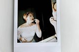 A polaroid of the author in her wedding dress, looking into a mirror and applying red lipstick