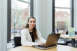 Katharine sat a desk in front of windows. She is using a laptop and smiling.