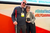 Two World Learning staff members (a man and a woman) stand side by side in front of a large backdrop in red and white. The man holds a walking cane, and the woman holds an award plaque.