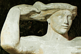 A close up view of the head and arms of a large statue. The statue’s face is tranquil, and the lines are drawn as in Ancient Egyptian statues. With one arm the figure reaches up to touch her hair, which unfurls in a way which suggests the surface of the sea.