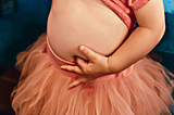 Picture of a toddler girl wearing a peach-colored tutu and top. Her top is pulled up, and her left index finger sticking into her navel.