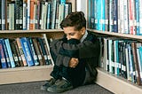 A young boy sits curled up in the corner of a library, hugging his knees to his chest with a worried or anxious expression.