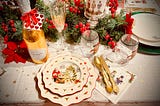A table laid out for a Christmas meal, complete with seasonal plates, sparkling champagne flutes, and plastic gold cutlery. There are also pine and holly garlands, poinsettias, a candle featuring nutcrackers, and a gold champagne bottle of chocolate con leche. The main place setting has dishes that feature a scene with Santa Claus, and there are some glasses with nutcrachers, as well. There is a tablecloth with gold stars on it.