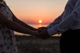 A couple faces each other holding hands with peaceful sunset in background.