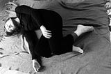 B/w photo of dancer lying on her side on a blanket, dressed in black, head resting on the floor, arms loose, legs bent.