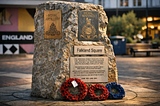 A granite memorial stone in Falkland Square, Poole, UK, displays plaques for the Royal Artillery and Royal Marines, with a dedication to local servicemen who died in the Falklands conflict. Two wreaths of red and blue poppies rest at its base. Warm evening light highlights the stone’s texture while the square and shops blur softly in the background, evoking quiet remembrance amid everyday life.