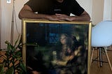 An artist sitting in a room and holding a framed painting.