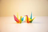 Two origami cranes made out of brightly covered paper sit on a table facing each other.