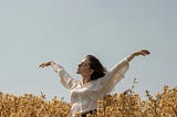 A woman stands in a flower field with her hands above her head.