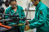 Two young women in teal safety jumpsuits work together in a machine shop.