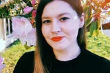 A photograph of Jenna James, a woman standing in front of a cherry blossom tree, wearing red lipstick and a black sweater.