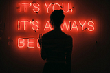A backlit young woman standing in front of a red neon sign that reads “it’s you, it’s always been…”
