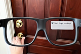 A first-person view through a pair of smart glasses, showing a hand reaching for a brass doorknob on a dark wooden door. A digital overlay in the upper right corner of the glasses’ display shows a notification that reads, “Don’t forget your keys!”