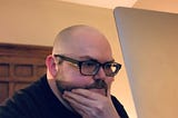 A bald person with glasses and a beard angsts looking at a laptop screen.