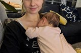 a woman smiles at the camera with a baby sleeping on her chest.