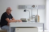 Still image of designer Dieter Rams sitting at a desk, surrounded by objects he has designed.