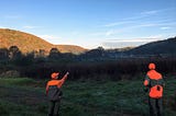Two men, wearing fluorescent orange hunting clothes, stand on a grass road. One points to a field as the sun rises over the valley they’re in.