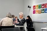 Two tables of women in discussion at a workshop. Both tables have three people at them. There is a display of the UN’s Sustainable Development Goals on the wall behind the tables.